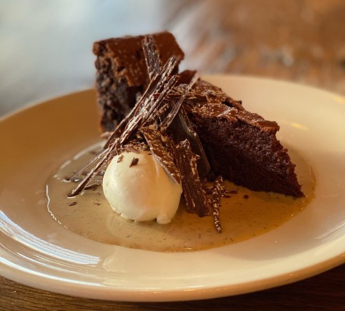 A slice of rich chocolate cake topped with chocolate shavings, served with a scoop of vanilla ice cream on a creamy sauce.