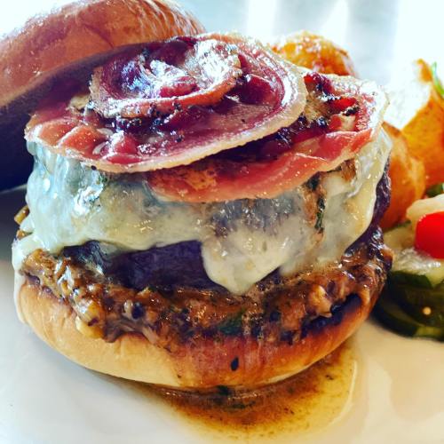 A gourmet burger topped with melted cheese, crispy bacon, and herbs, served with golden tater tots and pickled vegetables.