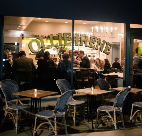 A cozy restaurant scene with patrons dining inside, visible through large windows, and outdoor seating with bistro chairs.