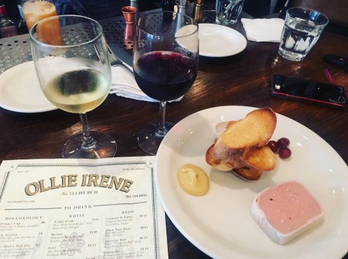 A table setting with two glasses of wine, a plate of pâté and toast, and a menu from Ollie Irene restaurant.