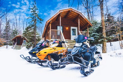 Three snowmobiles parked in front of a cozy log cabin surrounded by snow-covered trees under a blue sky.