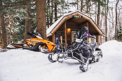 Two snowmobiles parked in front of a cozy cabin surrounded by snow-covered trees.