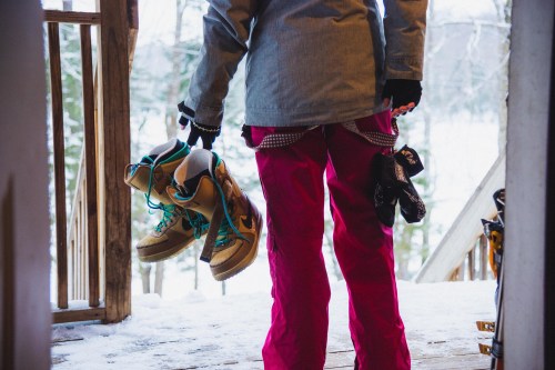 A person in pink pants holds two pairs of shoes while standing on a snowy porch. Trees are visible in the background.