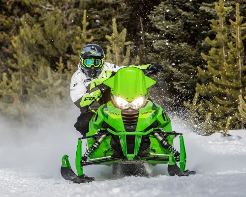A person riding a bright green snowmobile through snowy terrain, surrounded by trees.