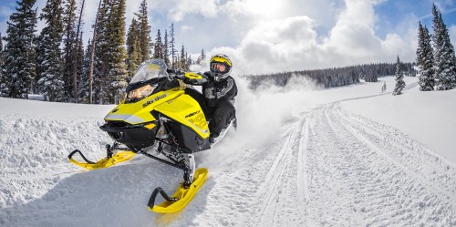 A person rides a bright yellow snowmobile through a snowy landscape, with trees and a blue sky in the background.