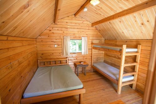 Cozy wooden cabin interior with a single bed, bunk bed, and a window, featuring warm wood paneling and natural light.