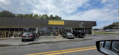 A storefront with large windows, displaying various items, and parked cars in front under a cloudy sky.