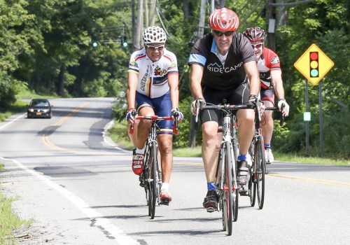 Three cyclists ride on a rural road, with trees lining the sides and a traffic signal in the background.