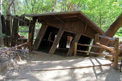 A tilted wooden structure surrounded by trees, with a sloped entrance and rustic fencing.