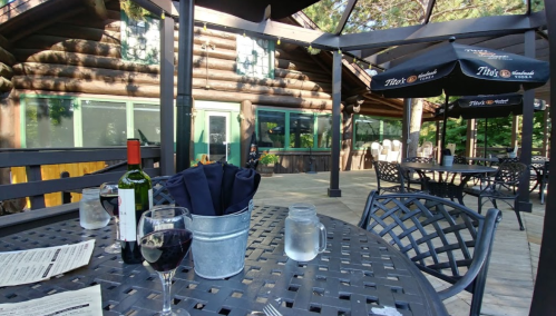Outdoor dining area with wooden logs, tables, wine, and a view of greenery under a shaded patio.
