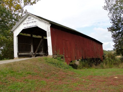 A red covered bridge from 1883, set on a grassy hill with trees in the background under a cloudy sky.