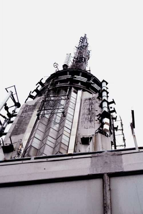 A tall communication tower with antennas and cables, viewed from below against a gray sky.