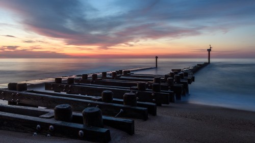 A serene pier extends into calm waters at sunset, with soft clouds and a colorful sky reflecting on the surface.