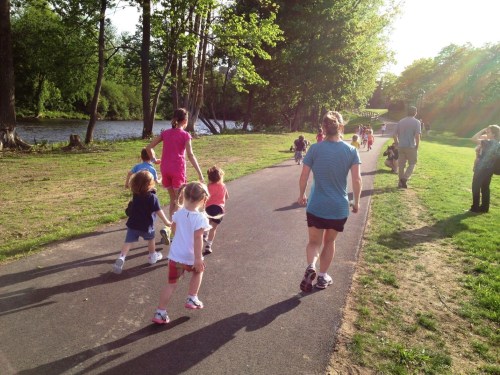 A group of children and adults walking along a path by a river, surrounded by trees and sunlight.