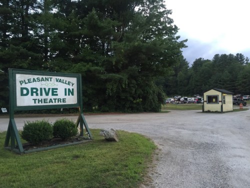Sign for Pleasant Valley Drive-In Theatre with a ticket booth and cars parked in the background, surrounded by trees.