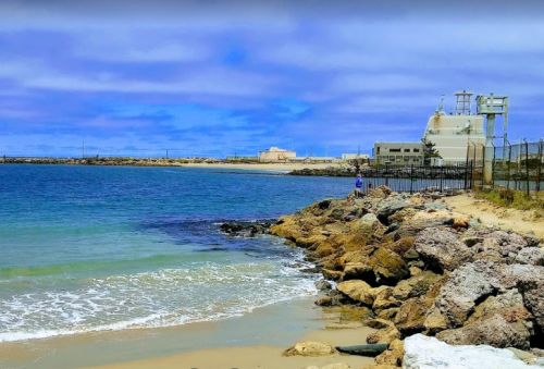 A rocky shoreline meets calm blue waters under a cloudy sky, with a building in the background.