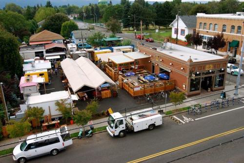 Aerial view of a food truck area with outdoor seating, surrounded by trees and buildings, and parked vehicles nearby.
