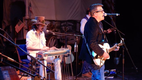 A band performs on stage, featuring a guitarist and a musician playing a steel guitar, with a backdrop of flags.