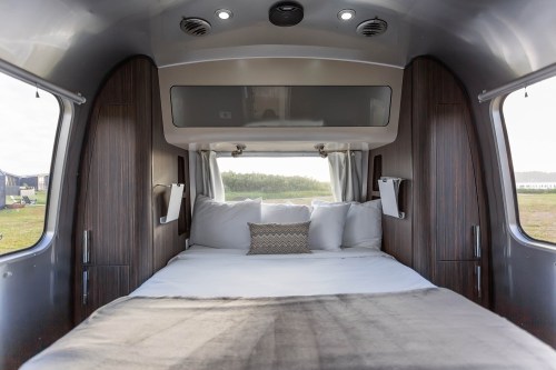 Cozy interior of a modern camper with a bed, decorative pillows, and large windows showcasing a green landscape outside.