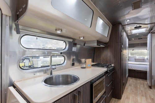 Modern kitchen in an Airstream trailer, featuring a sink, stove, and sleek cabinetry with large windows.