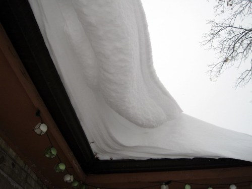 Snow accumulates on a roof, forming a large, curved overhang against a gray sky.