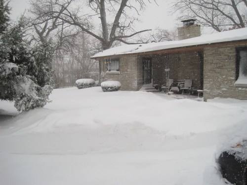 A snow-covered house with a stone exterior, surrounded by deep snowdrifts and bare trees in a winter landscape.