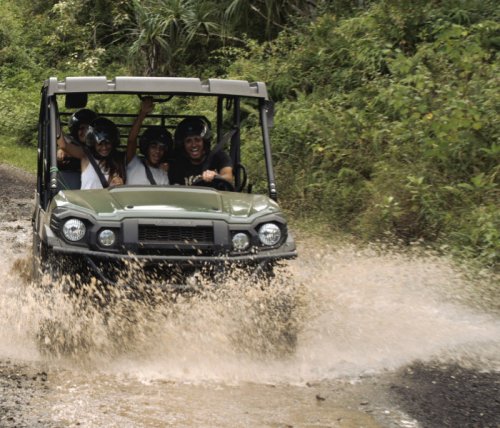A group of four people joyfully riding in an off-road vehicle splashing through water on a dirt path surrounded by greenery.