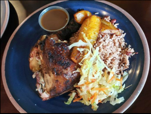 A plate of grilled chicken with rice, beans, fried plantains, and a side of salad, served with a small cup of sauce.