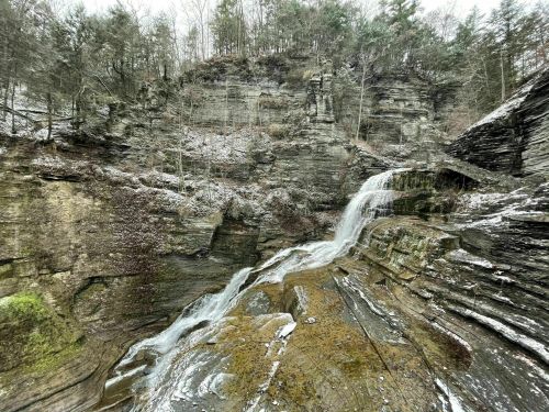 A serene waterfall cascades over rocky cliffs, surrounded by trees and a dusting of snow.