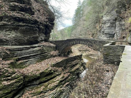 A stone bridge arches over a narrow creek, surrounded by steep, rocky cliffs and sparse trees in a serene landscape.