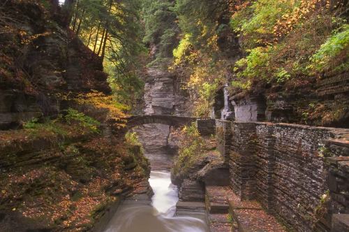 A serene canyon with a stone bridge, surrounded by colorful autumn foliage and flowing water.