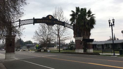 Archway sign reading "Historic Ripon" over a street, with palm trees and houses in the background on a cloudy day.