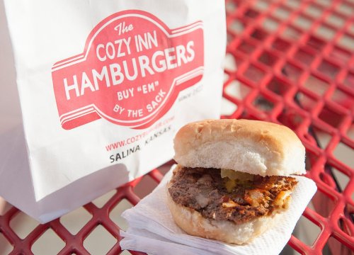 A cozy hamburger on a napkin next to a takeout bag from Cozy Inn Hamburgers, with a red table in the background.