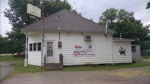 A small, white bar and grill building with a sign, surrounded by trees and gravel parking.
