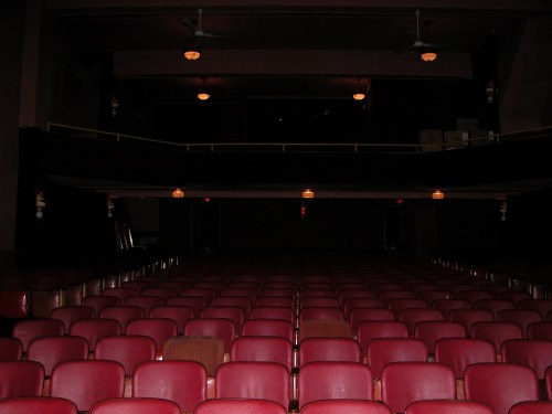 Empty theater with red seats and a dark stage, featuring overhead lights and a balcony in the background.