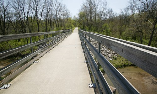 A concrete bridge with wooden railings spans a creek, surrounded by trees and greenery on a sunny day.