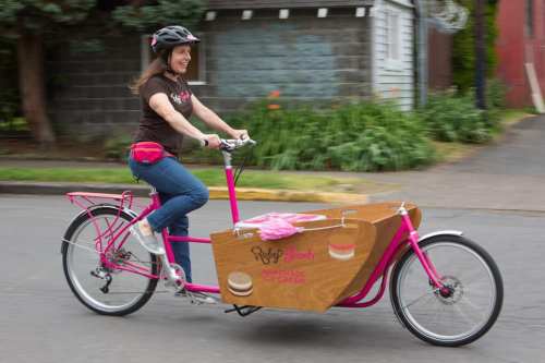 A woman rides a pink cargo bike with a wooden ice cream cart, smiling as she promotes Ruby Jewel ice cream.