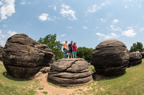 Three people stand on a large rock formation surrounded by spherical boulders under a blue sky with scattered clouds.