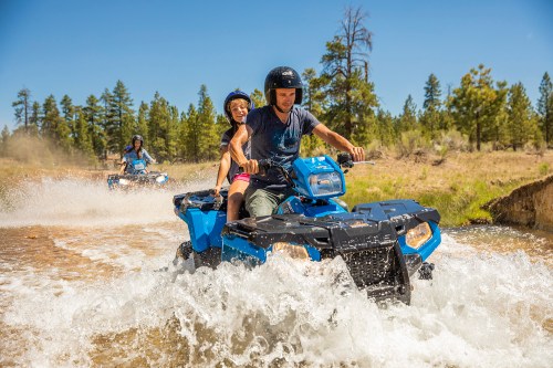 Two people ride an ATV through a shallow stream, splashing water, with trees in the background on a sunny day.