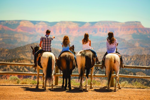 Four people on horseback overlook a scenic canyon landscape, with vibrant rock formations in the background.