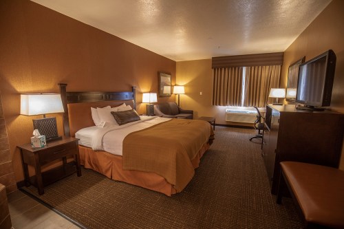 A cozy hotel room featuring a queen bed, armchair, desk, and a large window with curtains. Warm, inviting decor.