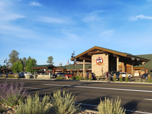 A rustic general store with a stone facade, surrounded by trees and parked cars, under a clear blue sky.