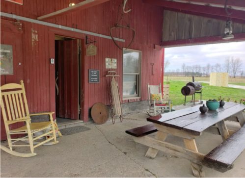A rustic outdoor seating area with a picnic table, rocking chairs, and a grill, set against a red barn backdrop.