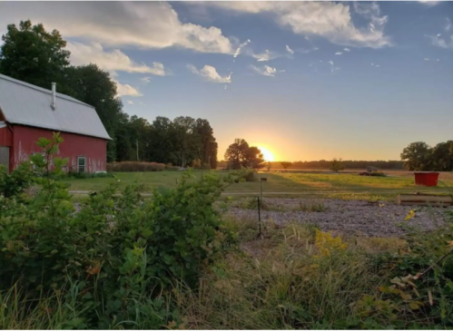 A red barn at sunset, surrounded by green fields and trees under a partly cloudy sky.
