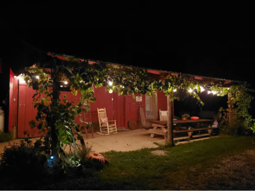 A cozy outdoor space at night with string lights, a rocking chair, and a picnic table surrounded by greenery.