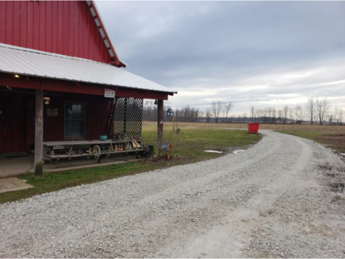 A red barn with a gravel path leading away, surrounded by open fields under a cloudy sky.