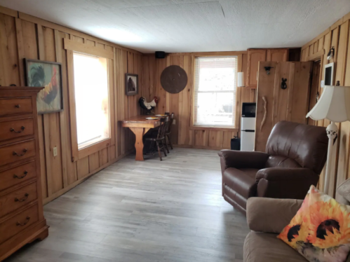 Cozy wooden interior with a dining table, chairs, a recliner, and a dresser, featuring natural light from large windows.