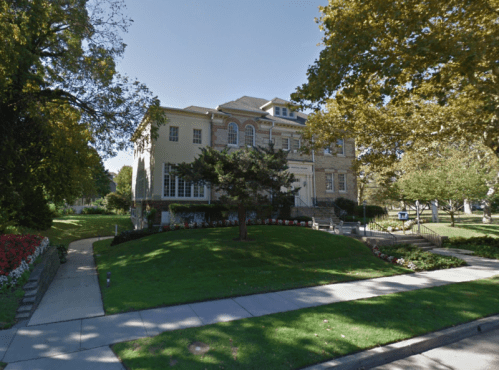 A large, historic building surrounded by trees and colorful flower beds on a sunny day.