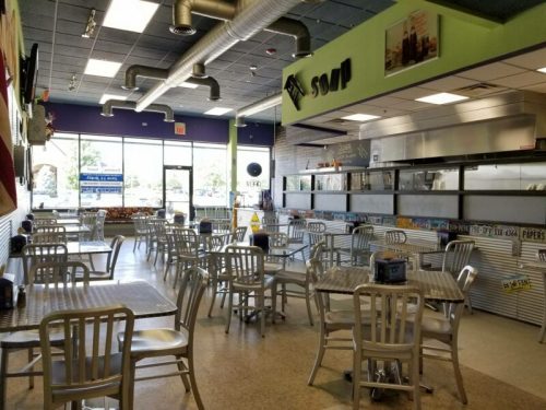 Interior of a casual dining restaurant with metal tables and chairs, large windows, and a counter in the background.