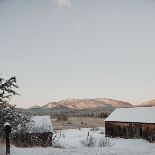 Snow-covered landscape with mountains in the background and rustic barns in the foreground. Clear winter sky.